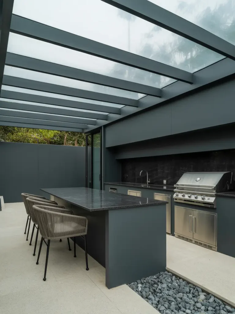 Modern weatherproof outdoor kitchen with sleek gray cabinetry, stainless steel grill, and a black countertop under a glass roof cover for all-weather protection.