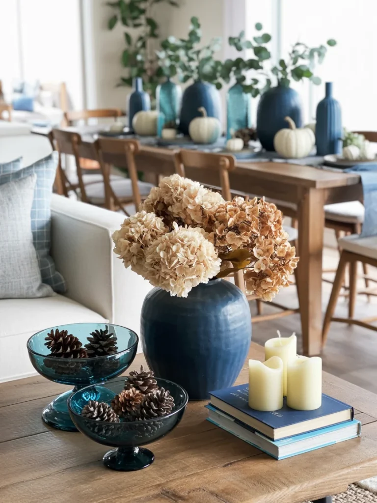 Blue fall centerpiece featuring navy vases with greenery, white pumpkins, dried hydrangeas, pinecones in glass bowls, and candles on a rustic wooden table.