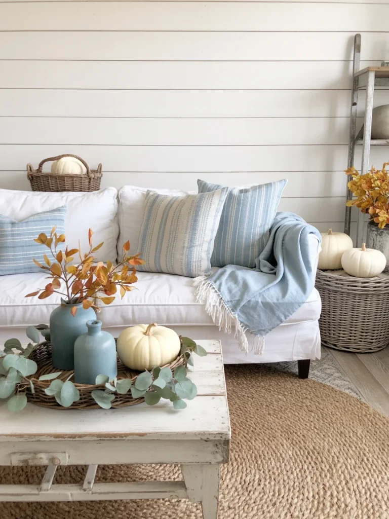Coastal fall living room with blue striped pillows, light blue throw, white pumpkins, and wicker accents styled with autumn branches.